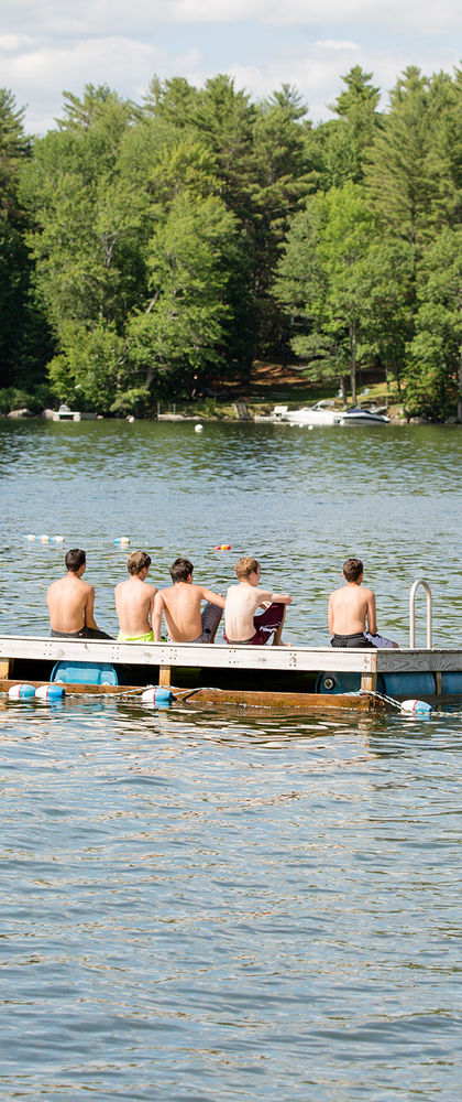 Five boys set on a floating dock in East Lake, looking back at Camp Manitou